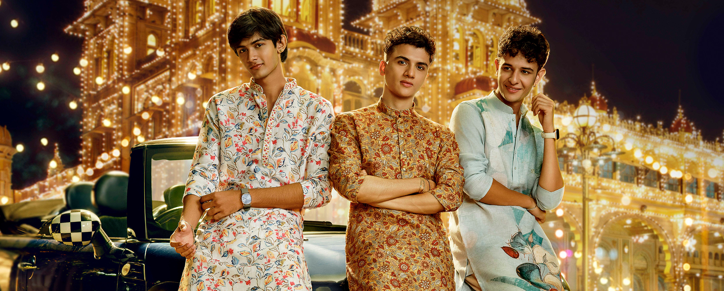 Young men in festive printed kurtas and accessories in front of an ornately lit palace backdrop.