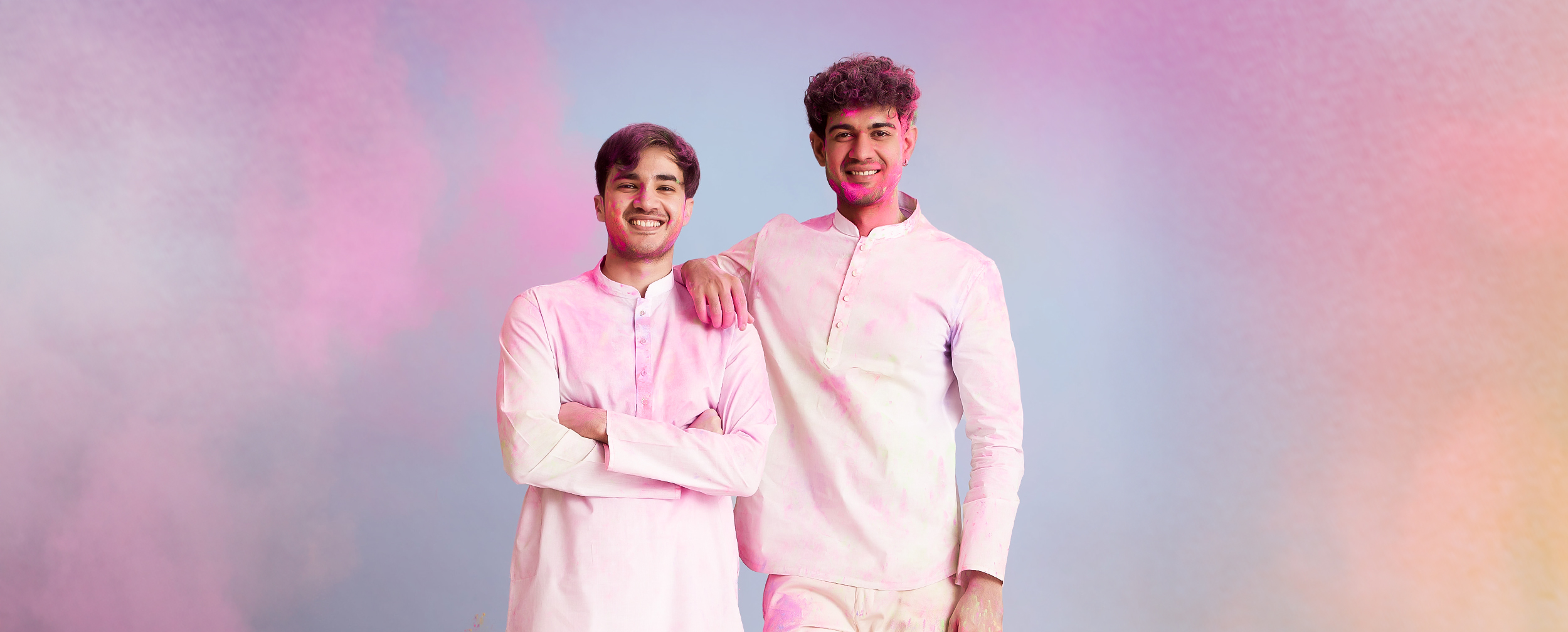 Two young men smiling in light white traditional kurta outfits, posing together against a colorful pastel Holi-themed background.