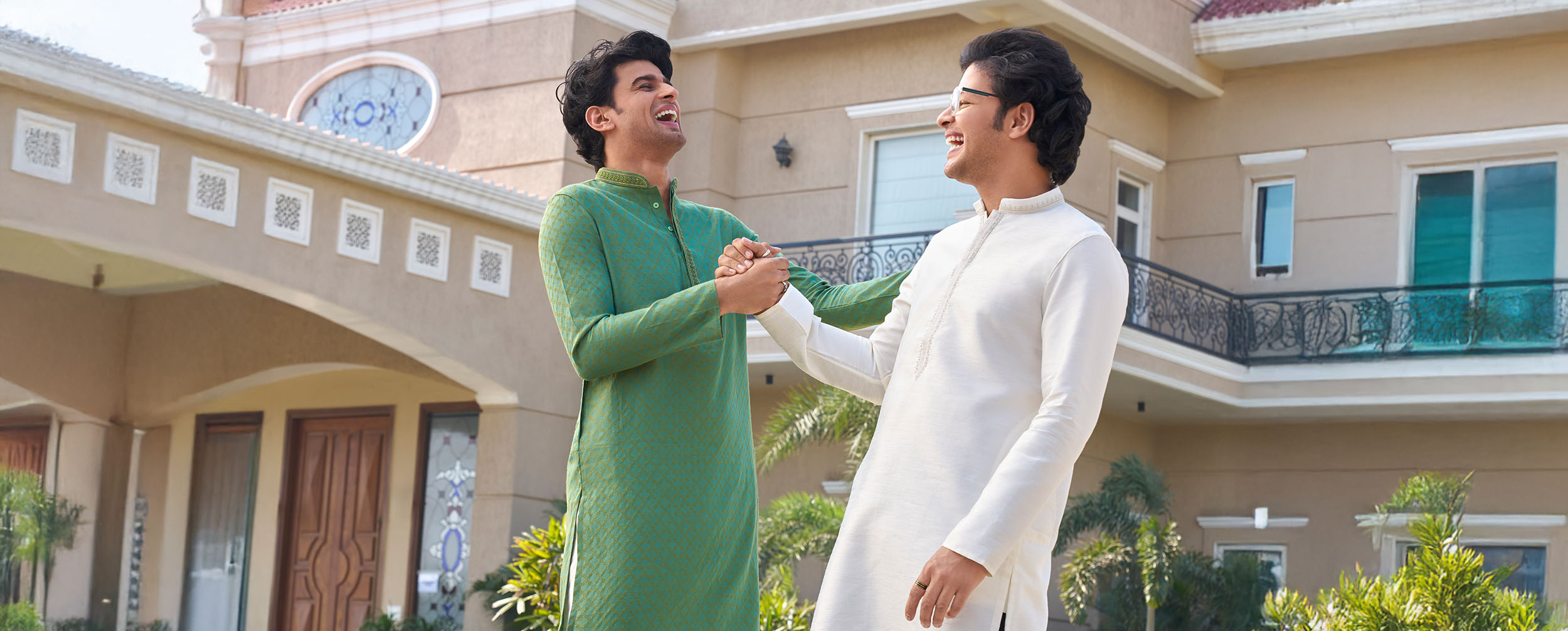 Two cheerful Indian men in traditional kurta pajamas, one in green and the other in white, greeting each other with joy outside a modern villa. Two cheerful Indian men in traditional kurta pajamas, one in green and the other in white, greeting each other with joy outside a modern villa.