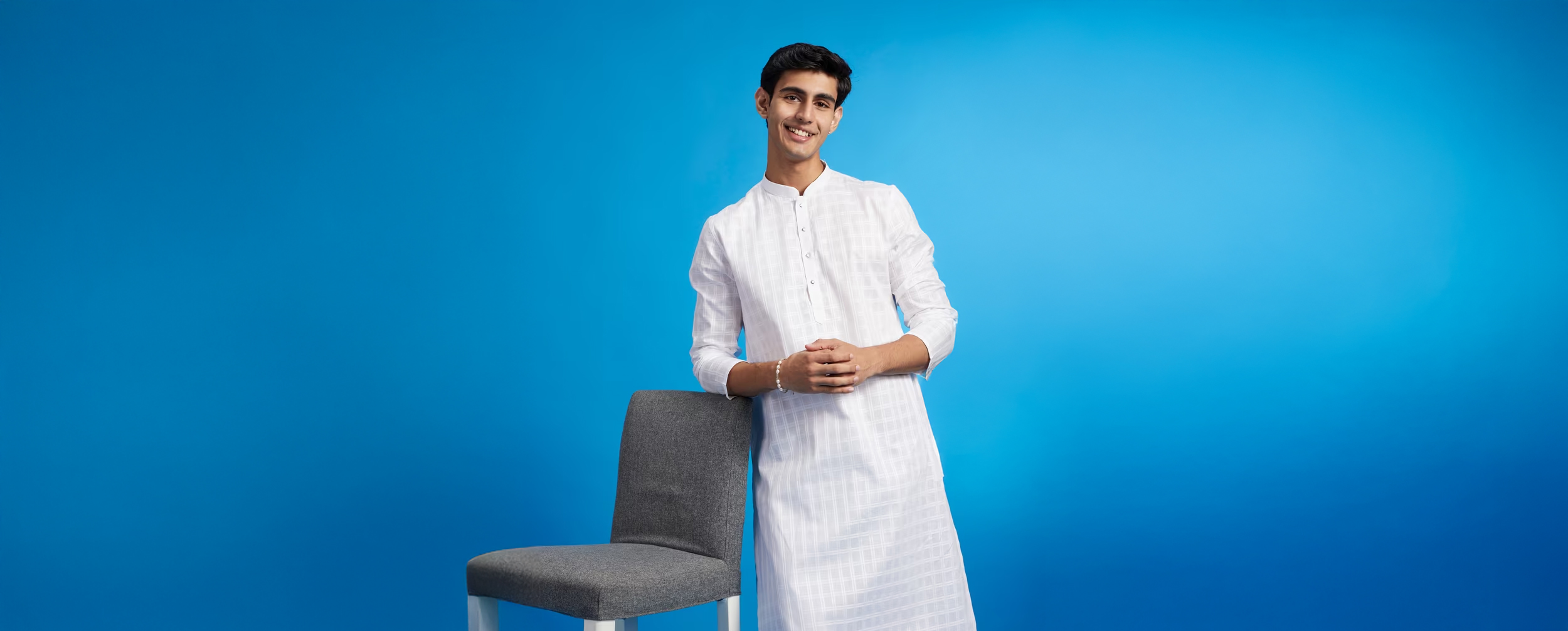 Young man in a white chikankari kurta, smiling and leaning casually against a grey chair, set against a bright blue background, showcasing traditional yet modern ethnic style. Young man in a white chikankari kurta, smiling and leaning casually against a grey chair, set against a bright blue background, showcasing traditional yet modern ethnic style.