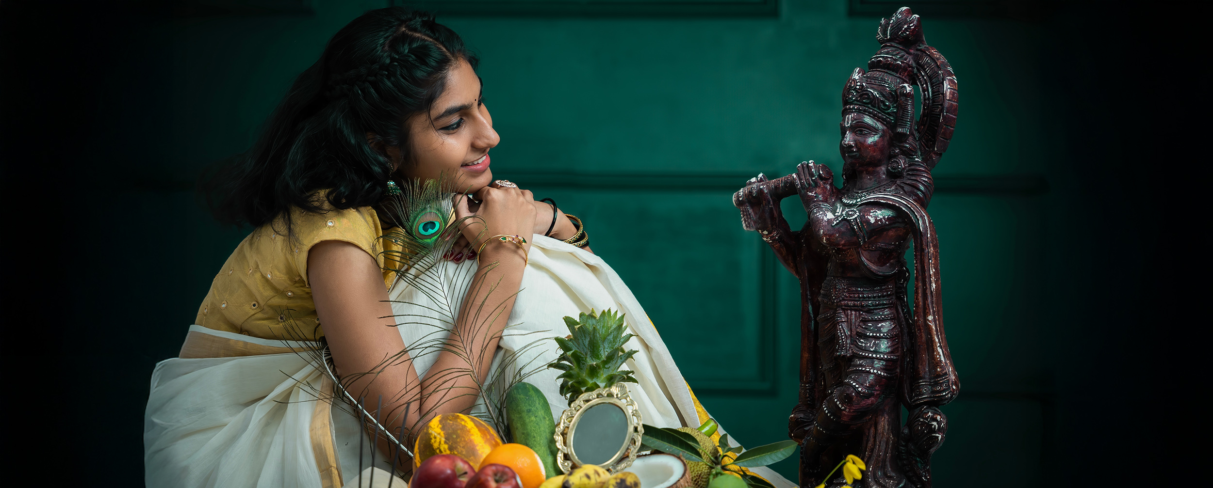Young woman in traditional attire smiling at a Lord Krishna idol, with fruits, peacock feather, mirror, and festive decorations arranged on a table during Janmashtami celebration. Young woman in traditional attire smiling at a Lord Krishna idol, with fruits, peacock feather, mirror, and festive decorations arranged on a table during Janmashtami celebration.