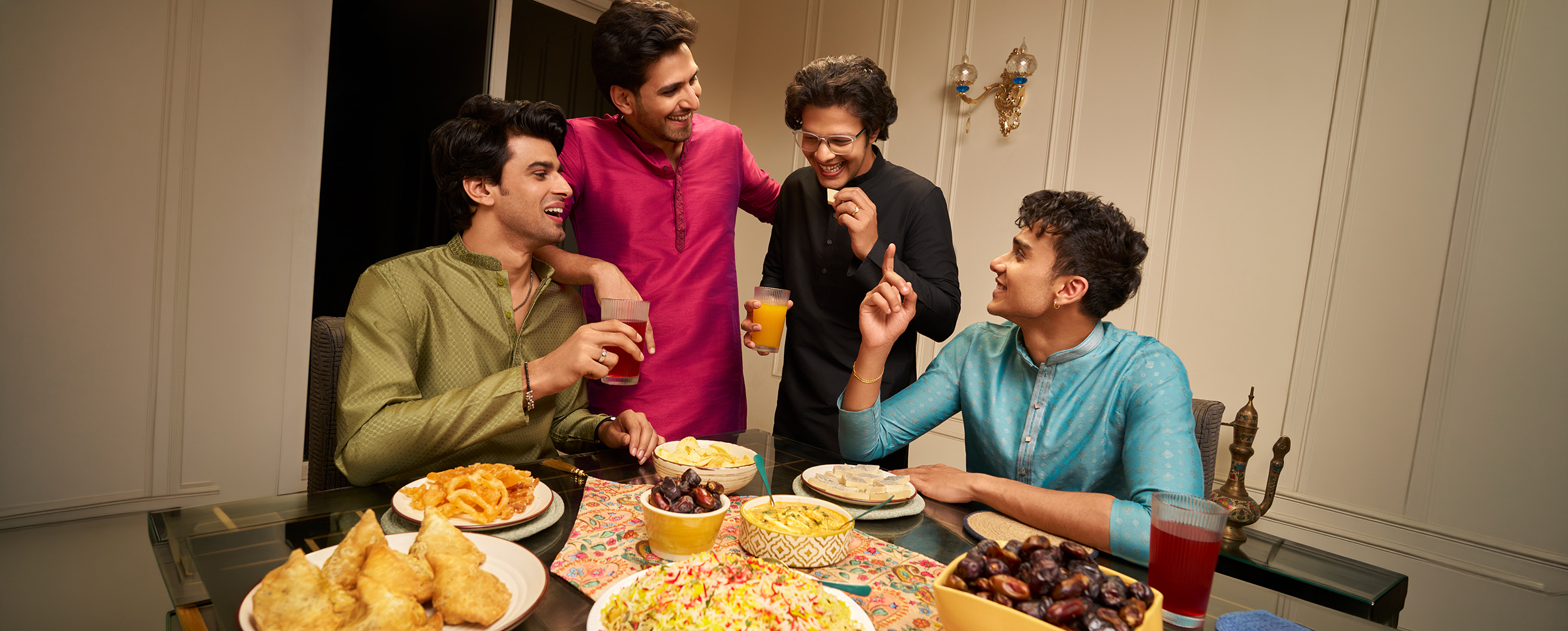 Friends in olive, pink, black, and teal kurtas enjoying snacks and drinks at a festive indoor gathering.