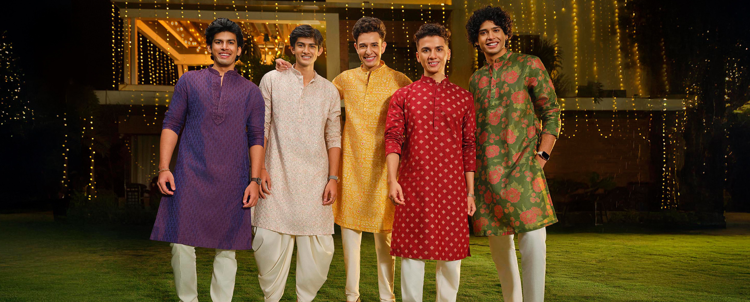Five men wearing colorful festive kurtas, standing together and smiling at an outdoor celebration.