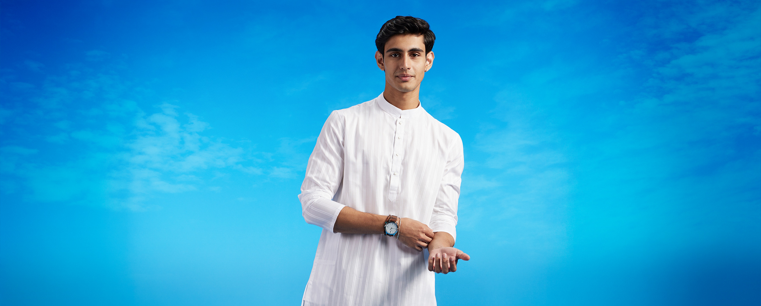 Man wearing a classic white kurta-pajama set, styled with a wristwatch, posing against a vibrant blue sky backdrop for modern Onam fashion inspiration. Man wearing a classic white kurta-pajama set, styled with a wristwatch, posing against a vibrant blue sky backdrop for modern Onam fashion inspiration.