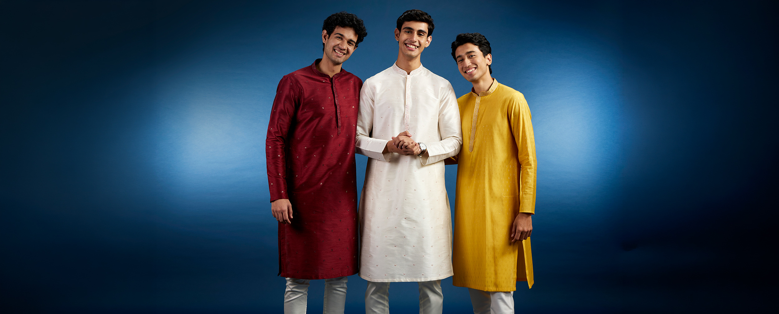 Three young men wearing solid-colored kurtas in red, ivory, and mustard demonstrate how clean silhouettes, proper length, and tailored fits create balanced proportions for shorter men.