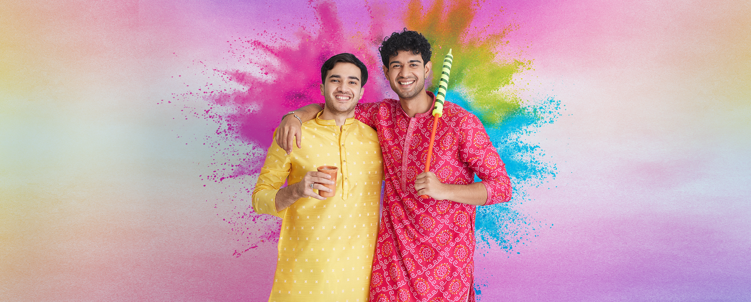 Two men in bright yellow and red kurtas smiling with arms around each other, holding Holi colors and a pichkari, with a vibrant color-splash background.