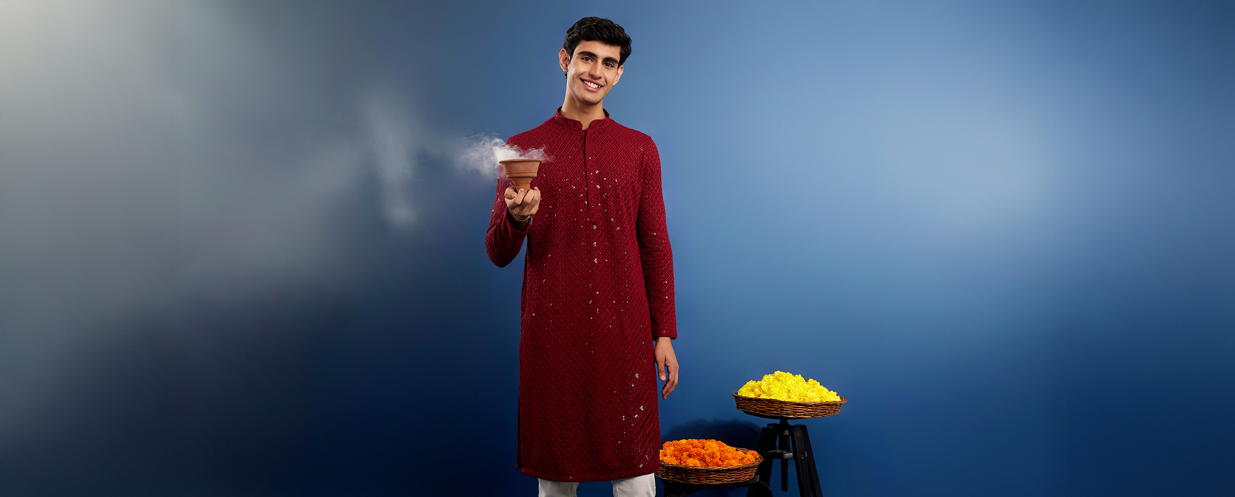 Man in maroon festive kurta with white pants holding incense pot, styled with marigold flowers for casual festive elegance
