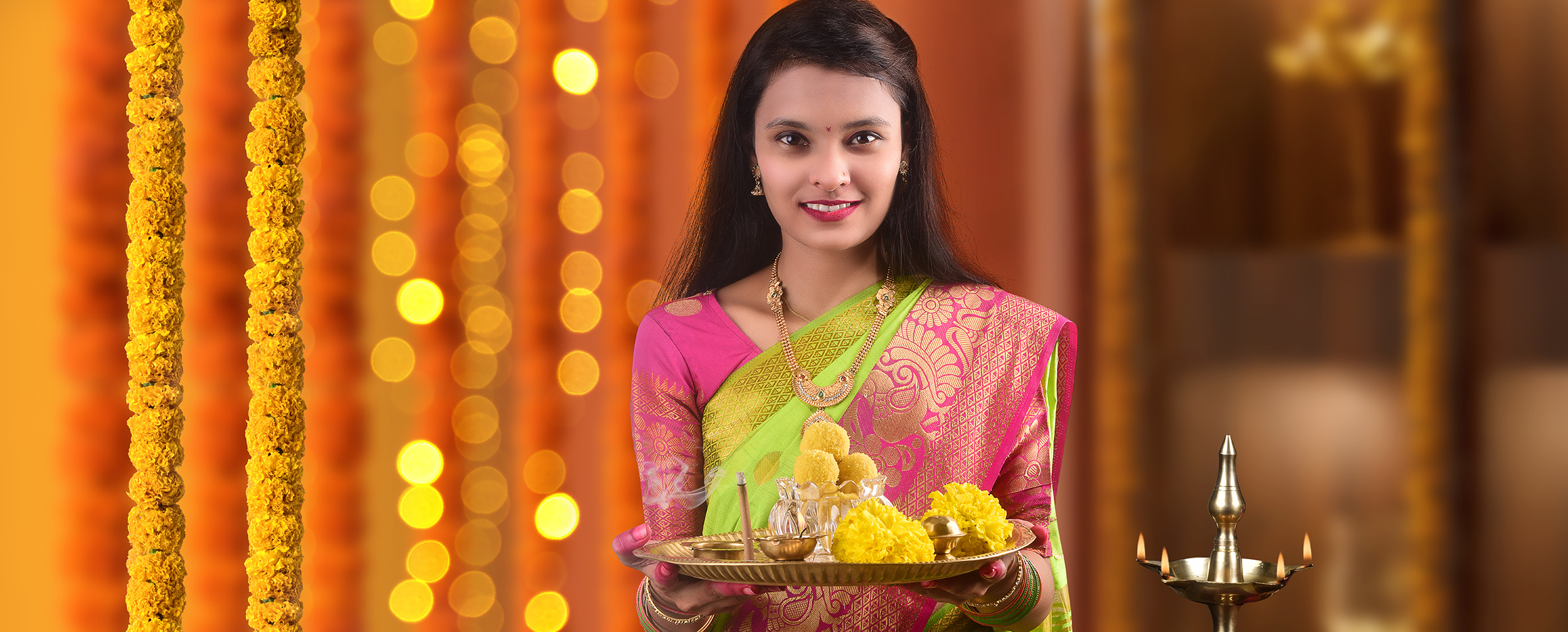 Woman in traditional saree holding decorated pooja thali with sweets, diya, and flowers during festive celebration