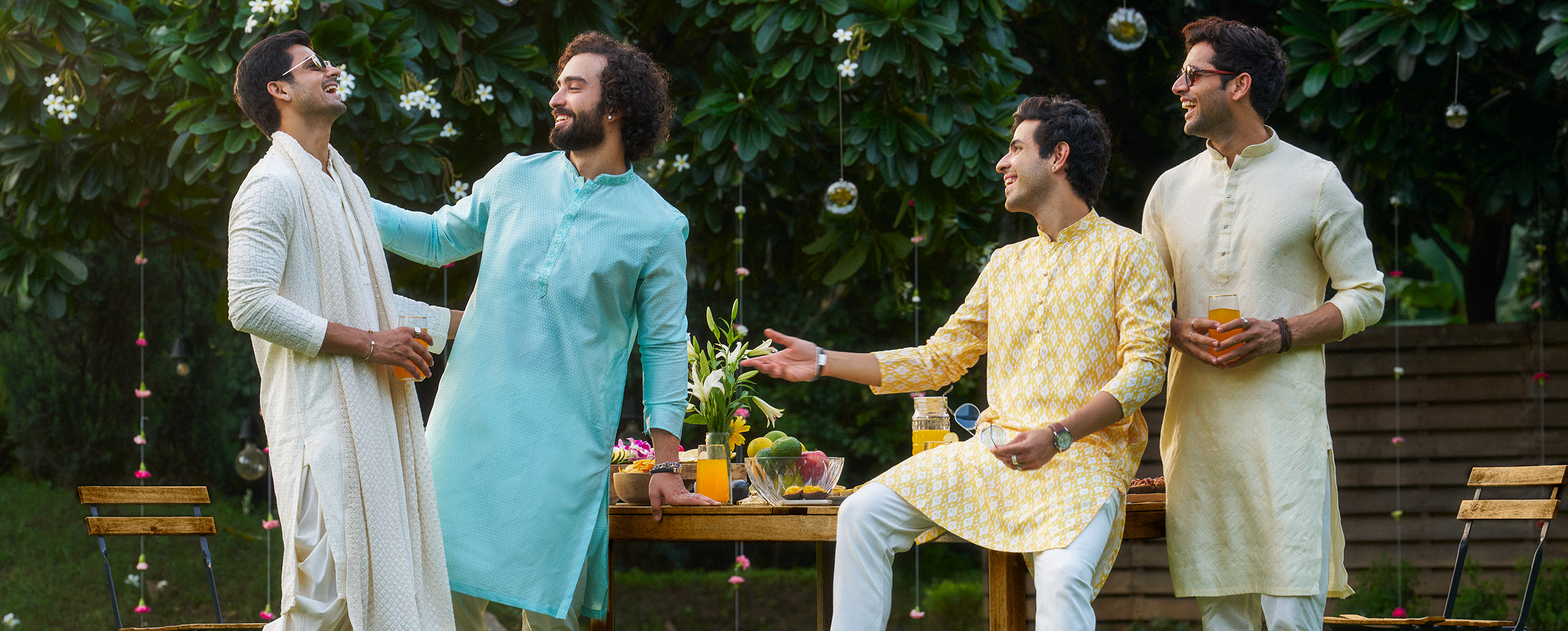 Four men in stylish pastel and bright cotton kurtas laughing and chatting outdoors at a summer gathering, with fresh drinks and fruits on a wooden table in a lush green garden. Four men in stylish pastel and bright cotton kurtas laughing and chatting outdoors at a summer gathering, with fresh drinks and fruits on a wooden table in a lush green garden.